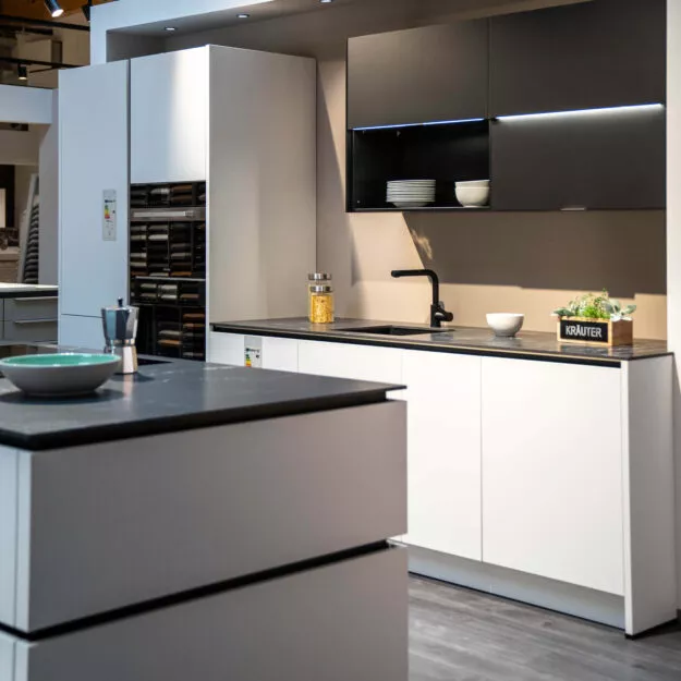 Moderne, weiße Küche mit dunkler Küchenarbeitsplatte und dunklen, beleuchteten Oberschränken und Kochinsel im Küchenstudio Tischer & Teuber.