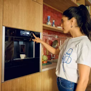 Frau bedient einen Neff Einbau-Kaffeevollautomaten in moderner Küche mit Holzfronten und roter Fliesenrückwand